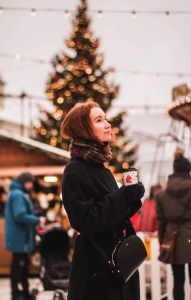 smiling woman having a hot drink in a Christmas market smiling woman looking up and holding a hot drink cup in a Christmas market with booths and a decorated Christmas tree in the background