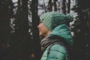 smiling girl in a green jacket, hat and a grey scarf smiling girl in the woods wearing a green puff jacket, green knitted hat and a grey scarf